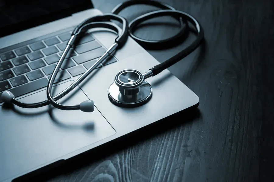 Telemedicine-Doctor An open laptop with a stethoscope lying on it on a wooden table, symbolizing telemedicine care from Julie Kane, NP in Newfoundland.